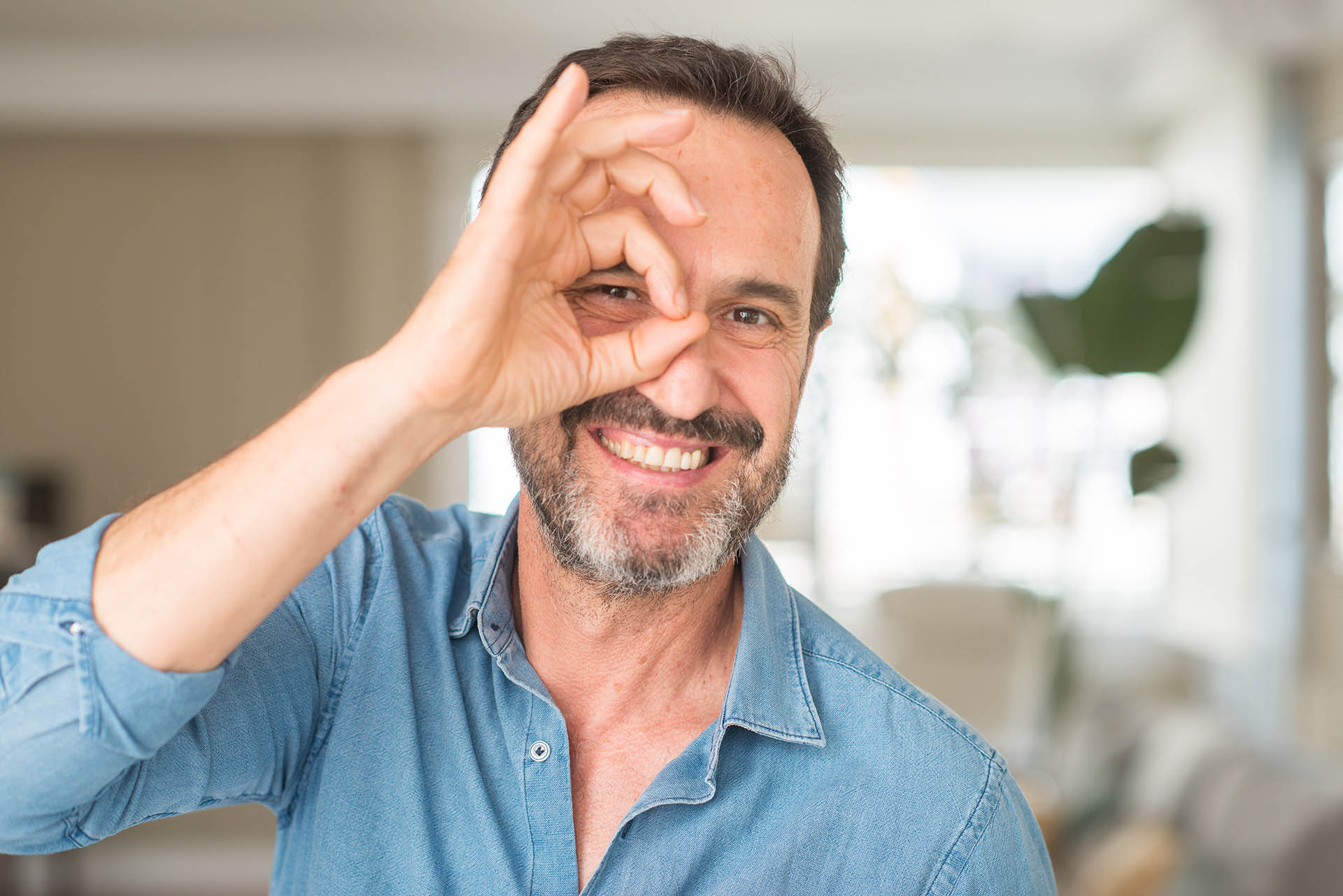 Handsome middle age man with happy face smiling doing ok sign with hand on eye looking through fingers